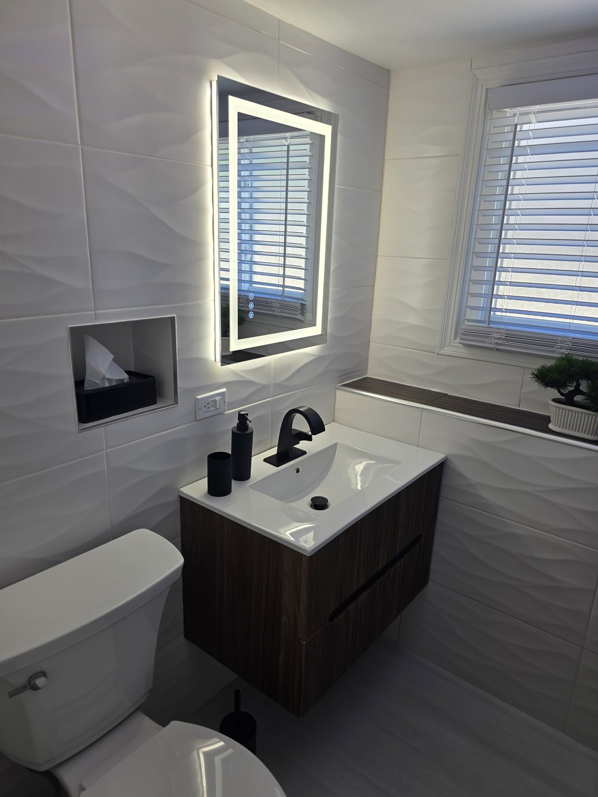 After: the same bathroom finished with wave-pattern white wall tile, a backlit mirror and a floating dark wood-grain vanity with a black faucet